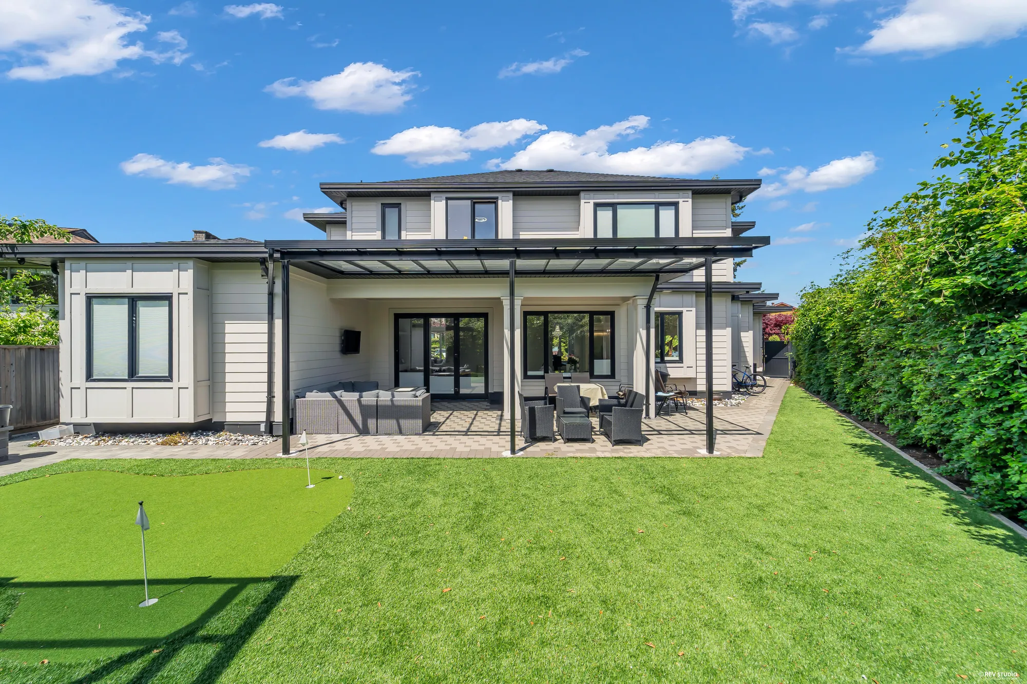 The back of the house from the end of the yard: a covered patio under a glass-roofed canopy, outdoor sofas and a table and chairs, a lawn with a putting green in the foreground, a tall hedge at the right