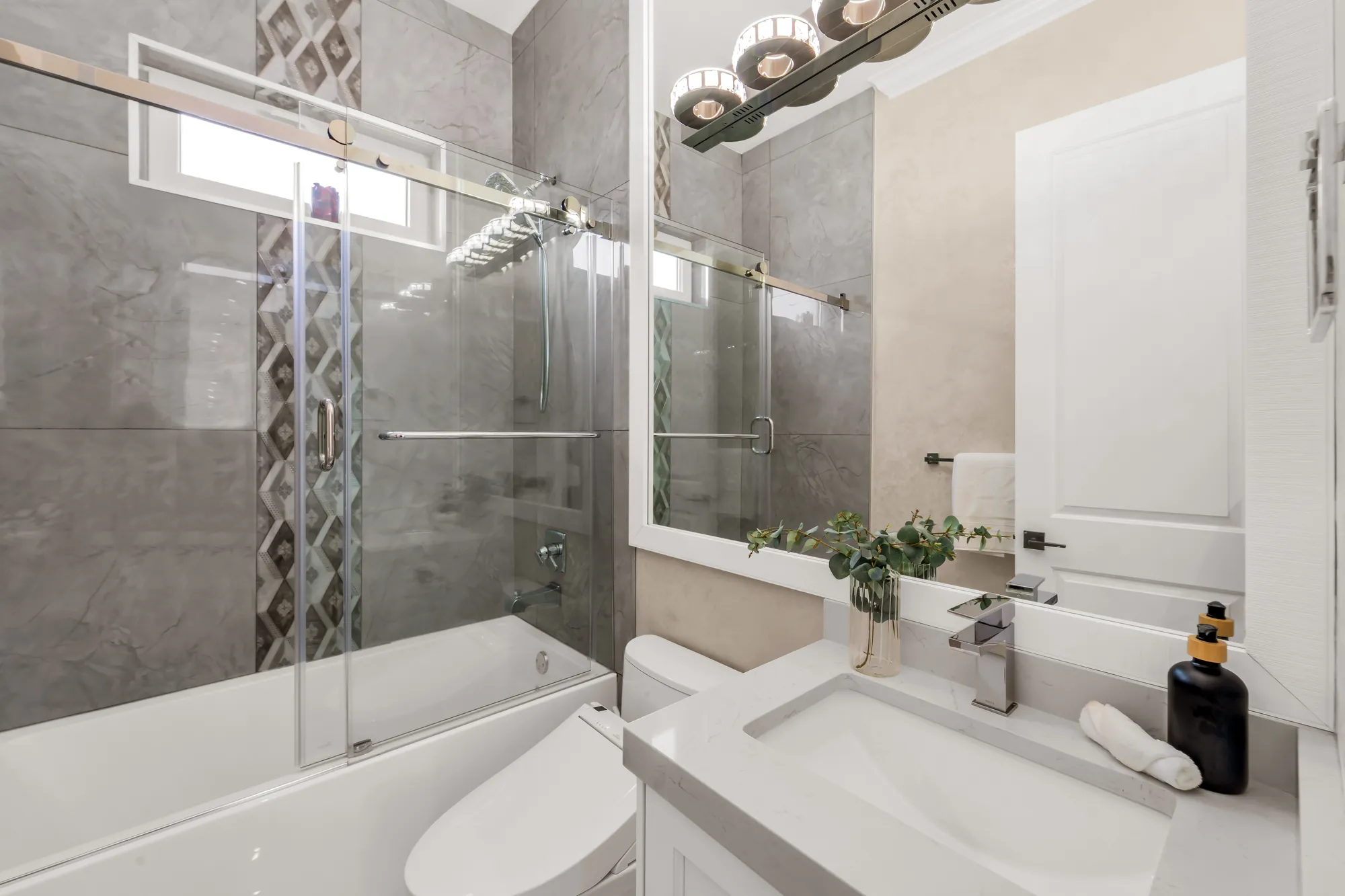 A bathroom on the main floor: a tub behind a sliding glass door, grey tile with a vertical mosaic strip, a white vanity, a toilet, a row of globe lights above the mirror