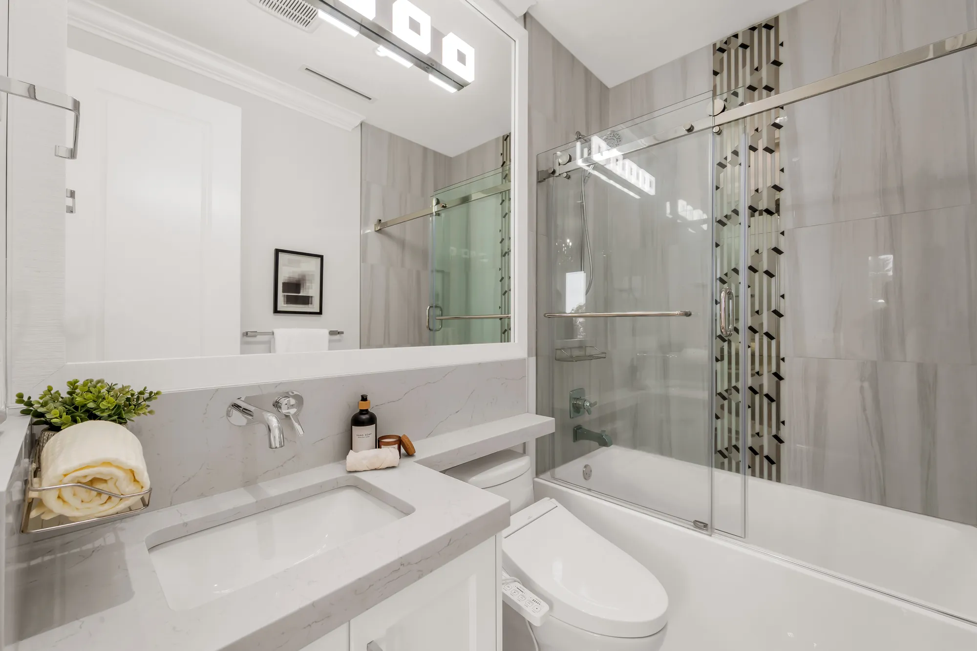 A bathroom with a tub behind a sliding glass door, grey tile with a vertical black and white mosaic strip, a white vanity with a wall-mounted faucet, a toilet