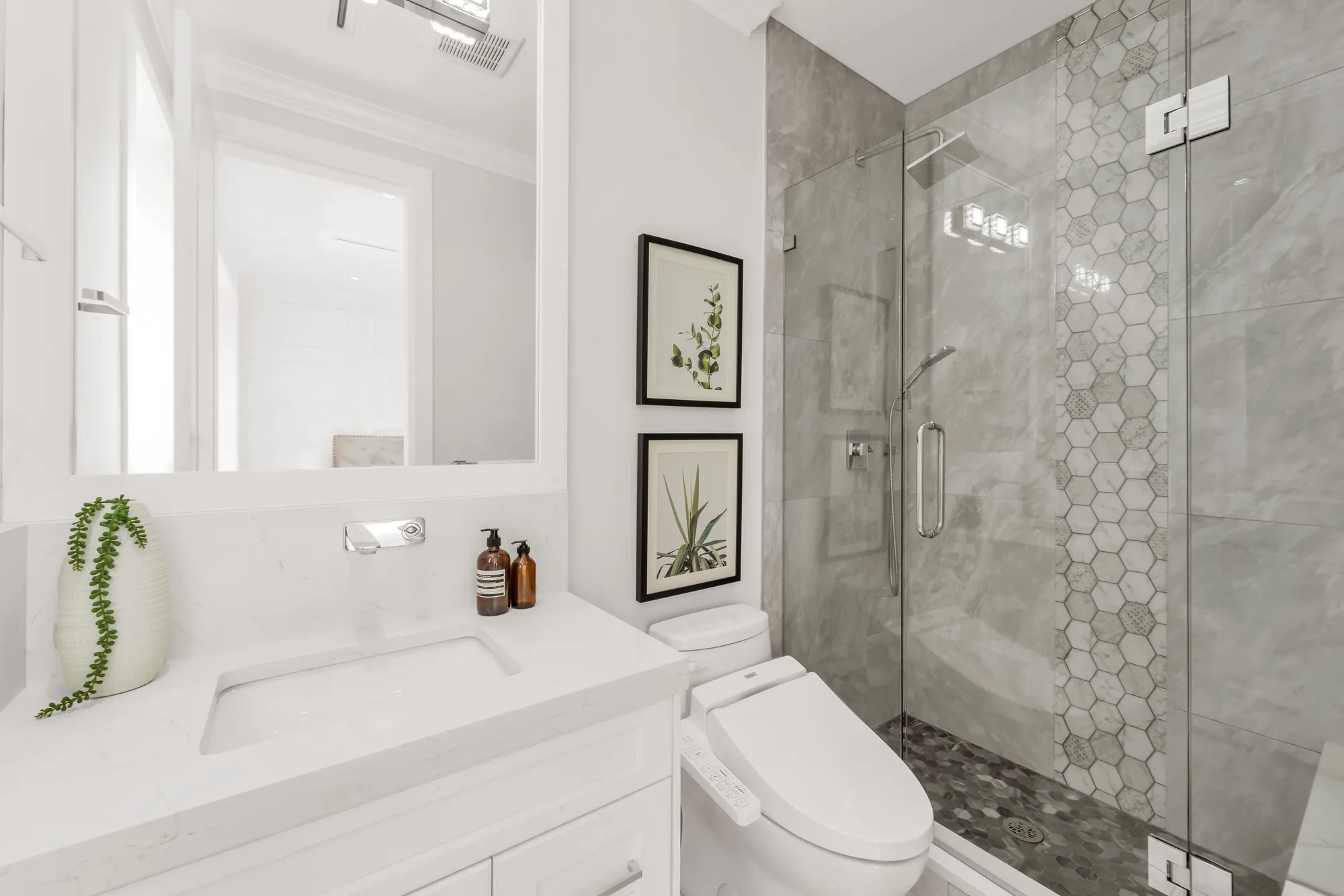 A bathroom with a glass-enclosed shower, a hexagon mosaic strip, a white vanity, a toilet, two framed botanical prints