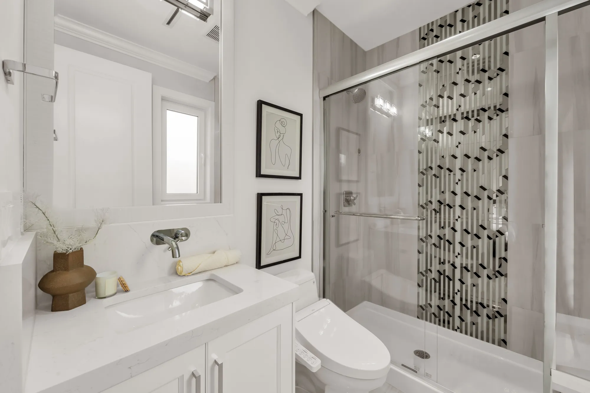 A bathroom with a shower behind a sliding glass door, a vertical black and white mosaic strip, a white vanity, a toilet, two line drawings