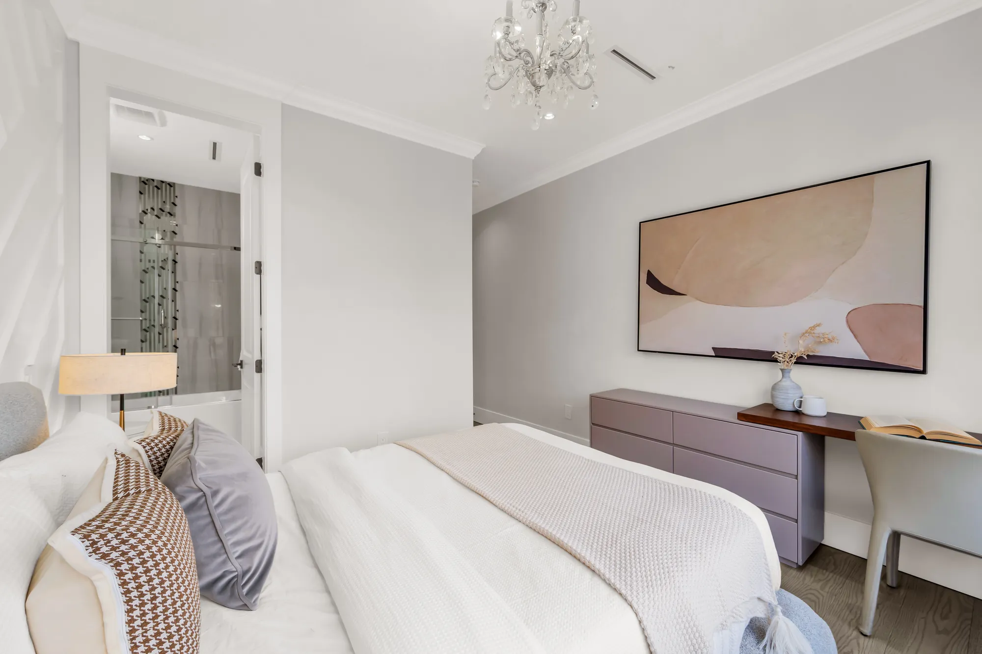 The same bedroom from the window side: the bed, a lilac dresser with a desk, an abstract painting, a bathroom through the door at the left