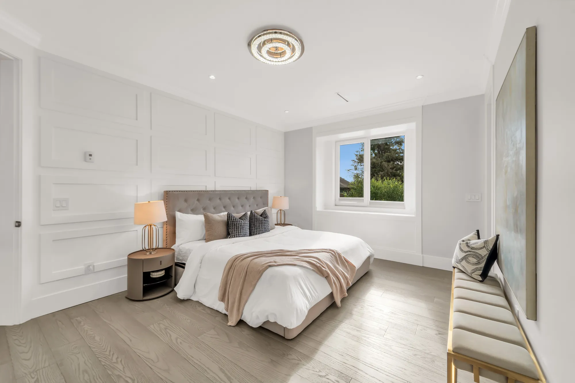 A bedroom with panelled walls: a bed with a grey tufted headboard, a window, a bench with brass legs, a painting