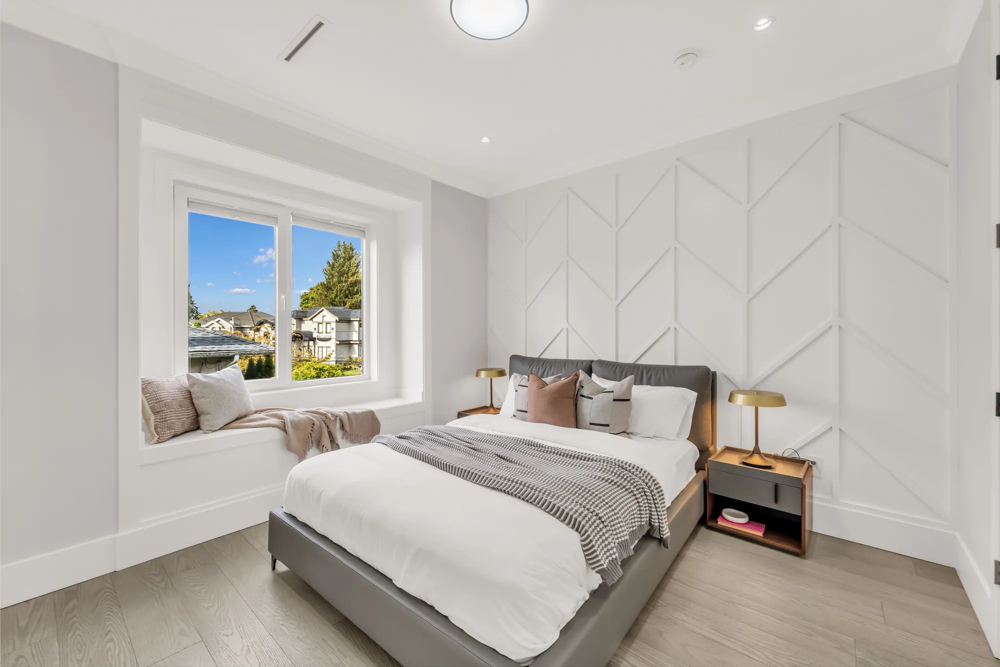 A bedroom with a white chevron-panelled wall and a window seat, a bed with a grey headboard, night tables and brass lamps