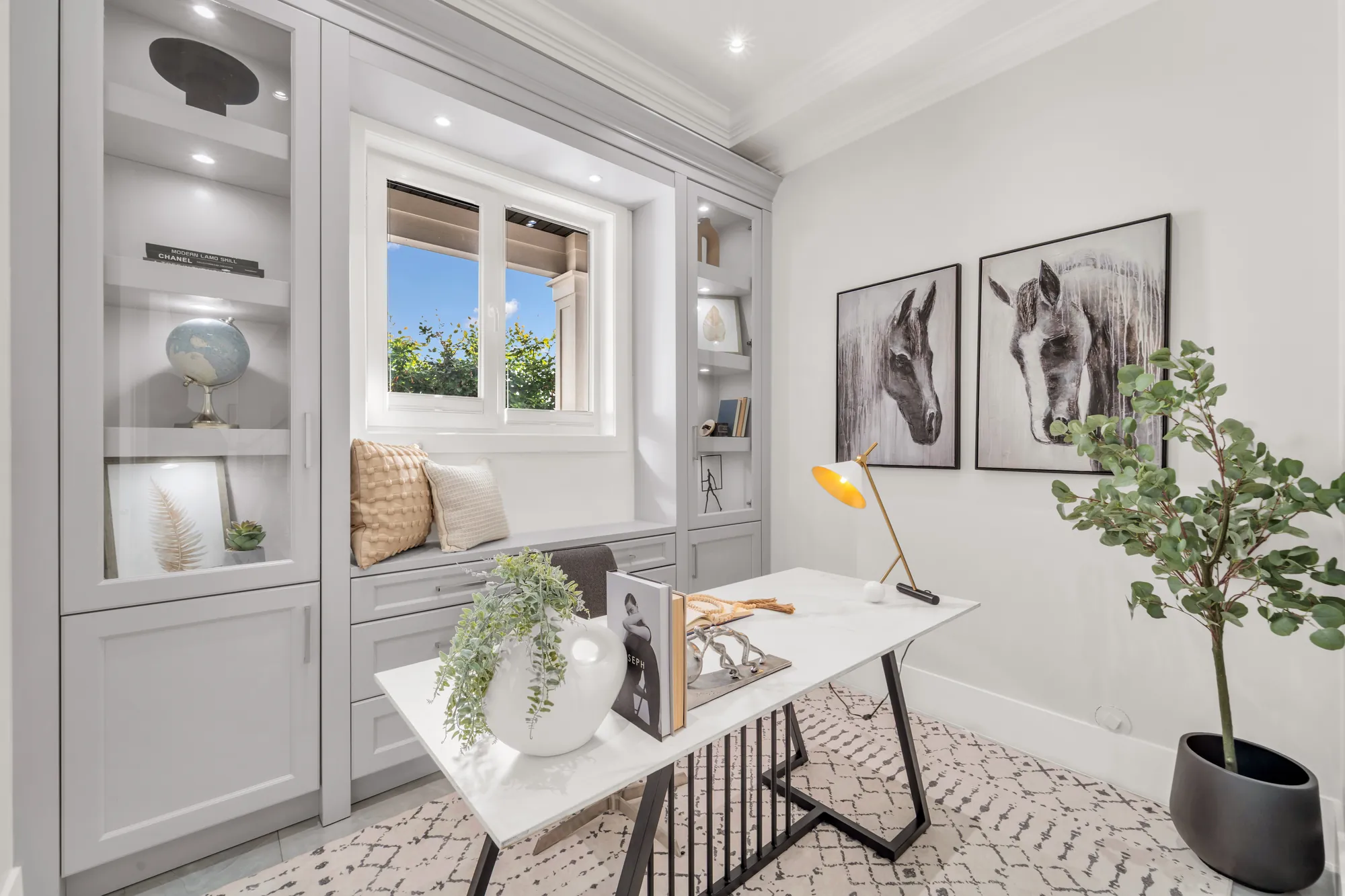 The den: built-in shelving with lighting, a window seat under a window, a desk and a lamp, two framed horse prints, a potted tree