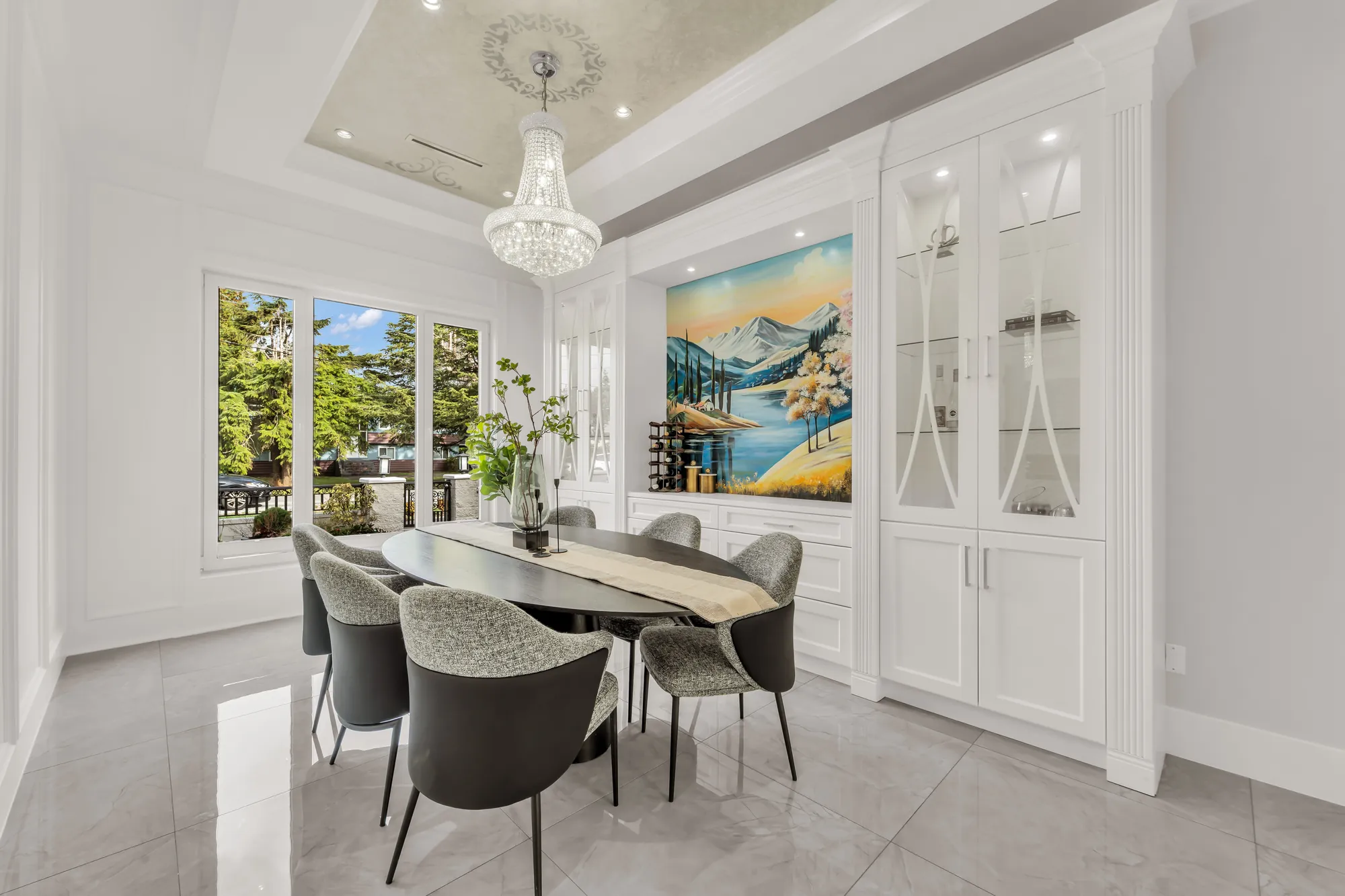 The dining room: an oval table and six upholstered chairs under a crystal chandelier in a tray ceiling, a large landscape painting, built-in glass-front cabinets, windows onto the front garden