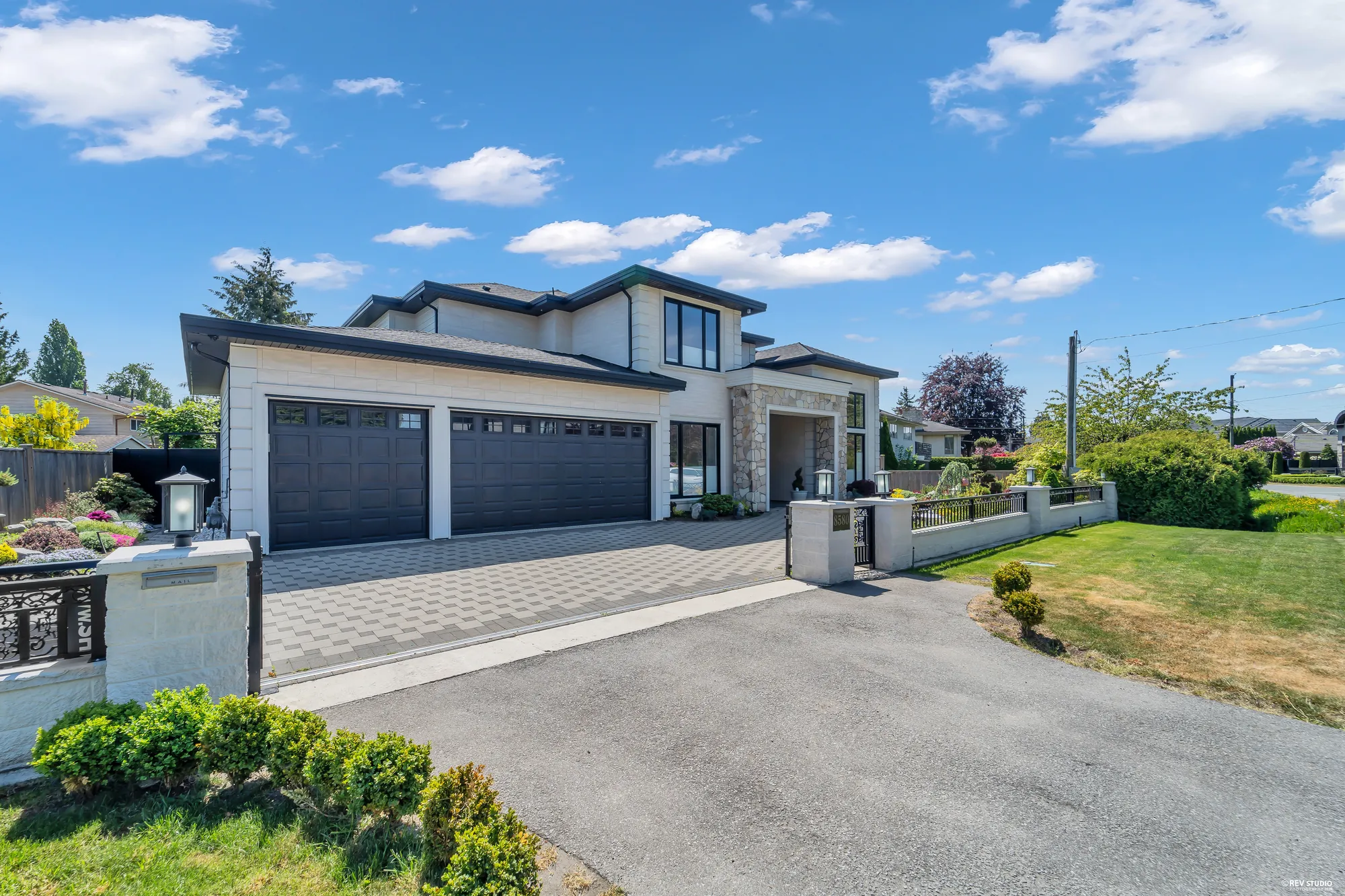 The drive from the street: a paved apron and a driveway in grey pavers to the garage doors, the white wall and railing along the front garden, the lawn at the right, clipped shrubs in the foreground