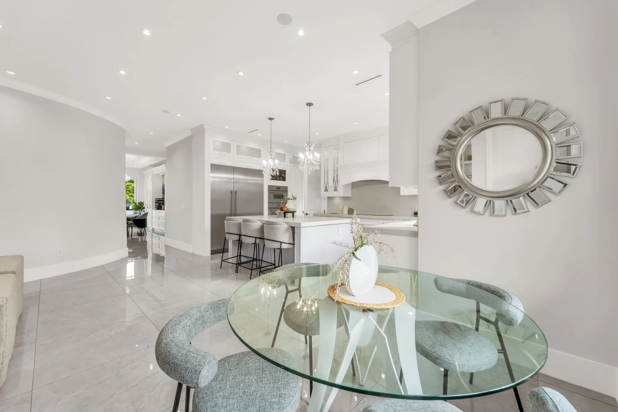 The eating area: a round glass table and four upholstered chairs, a sunburst mirror on the wall, the kitchen island and the chandeliers beyond