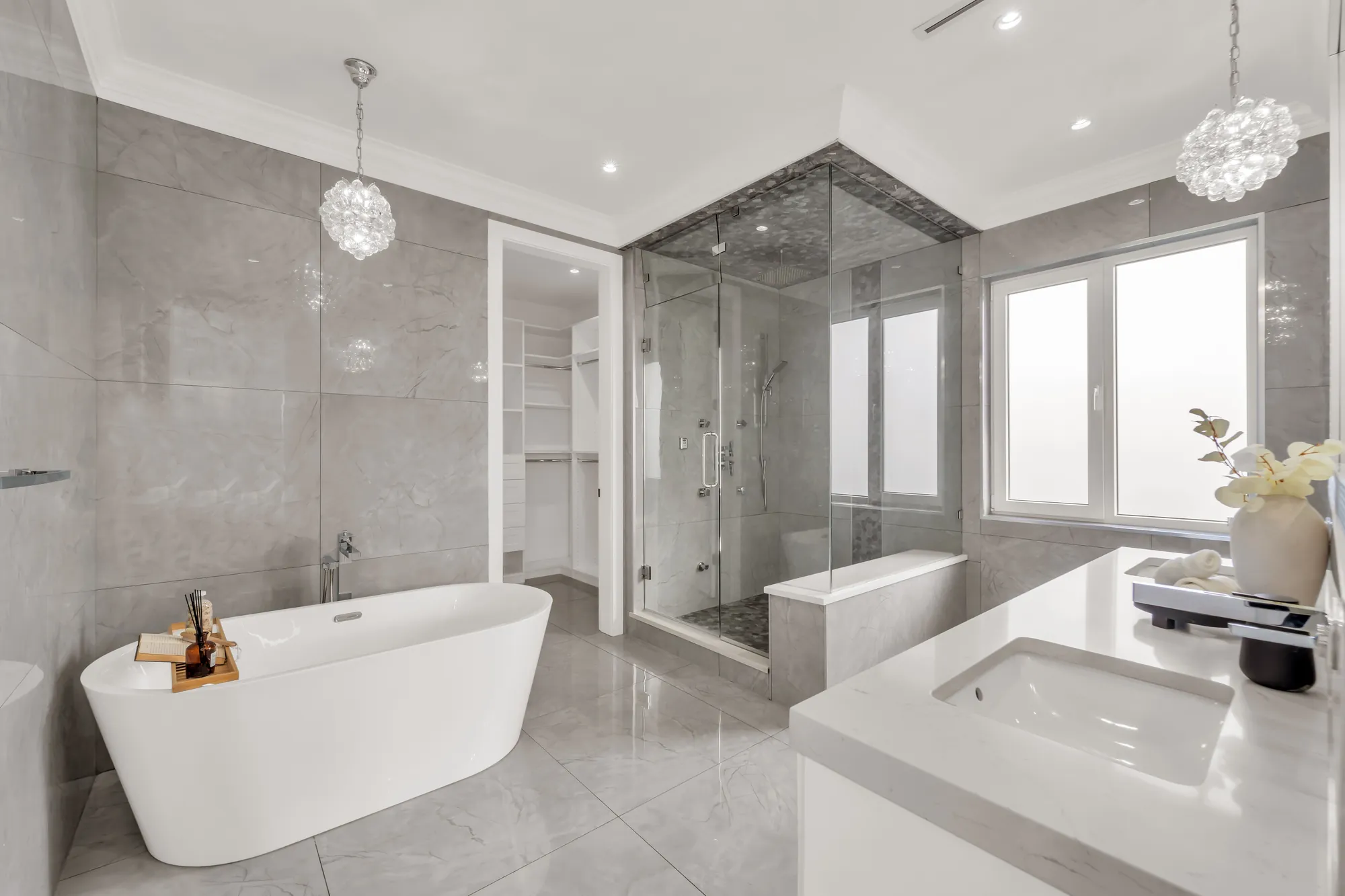 The primary bathroom: a freestanding tub, a glass-enclosed shower with a bench, a white vanity, large grey tiles, crystal pendants, a frosted window