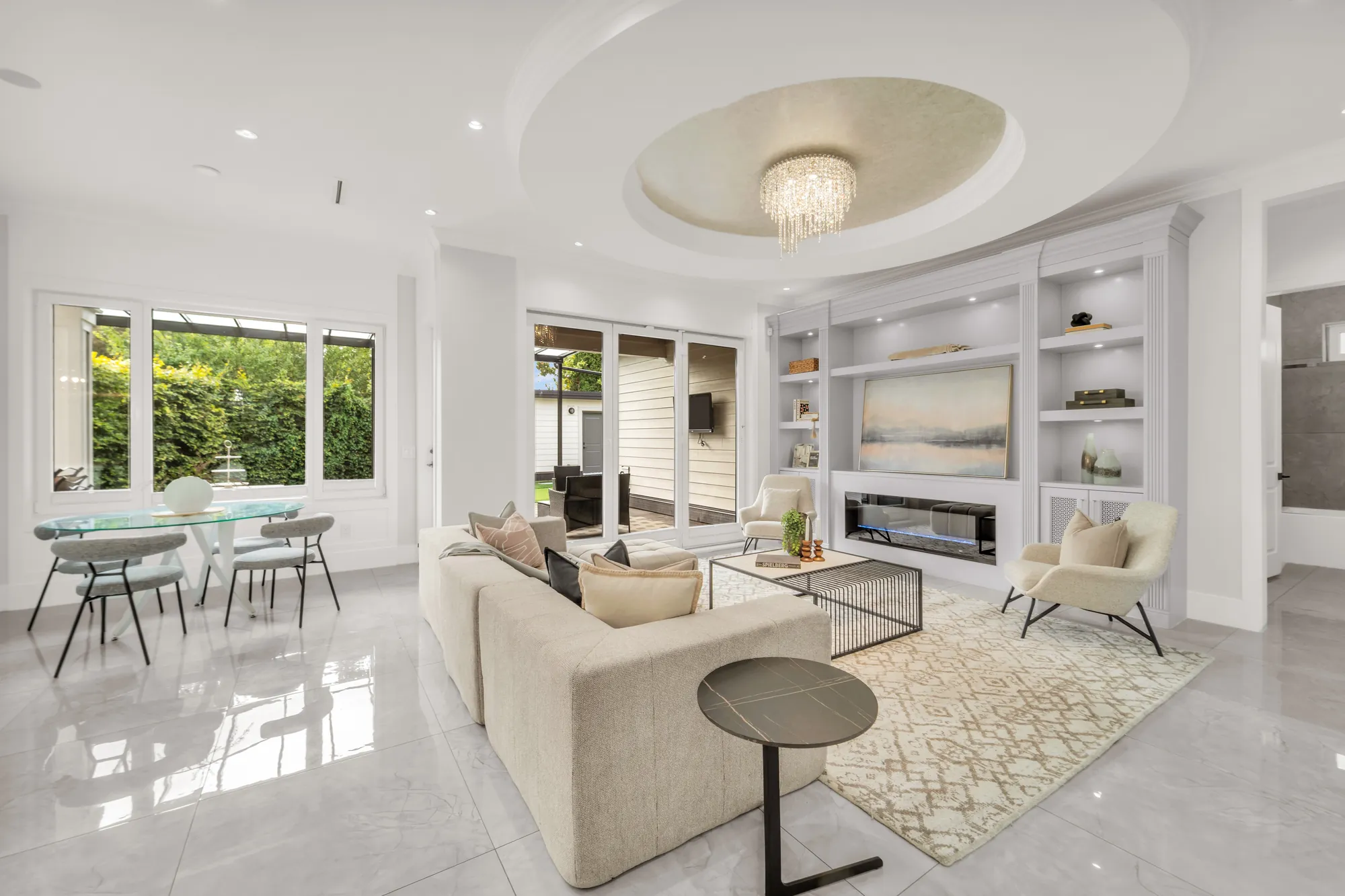 The family room: a round tray ceiling with a crystal light, a wall of built-in shelving around an electric fireplace and a painting, a sectional sofa and lounge chairs on a patterned rug, the eating area with a glass table at the left, windows onto the covered patio