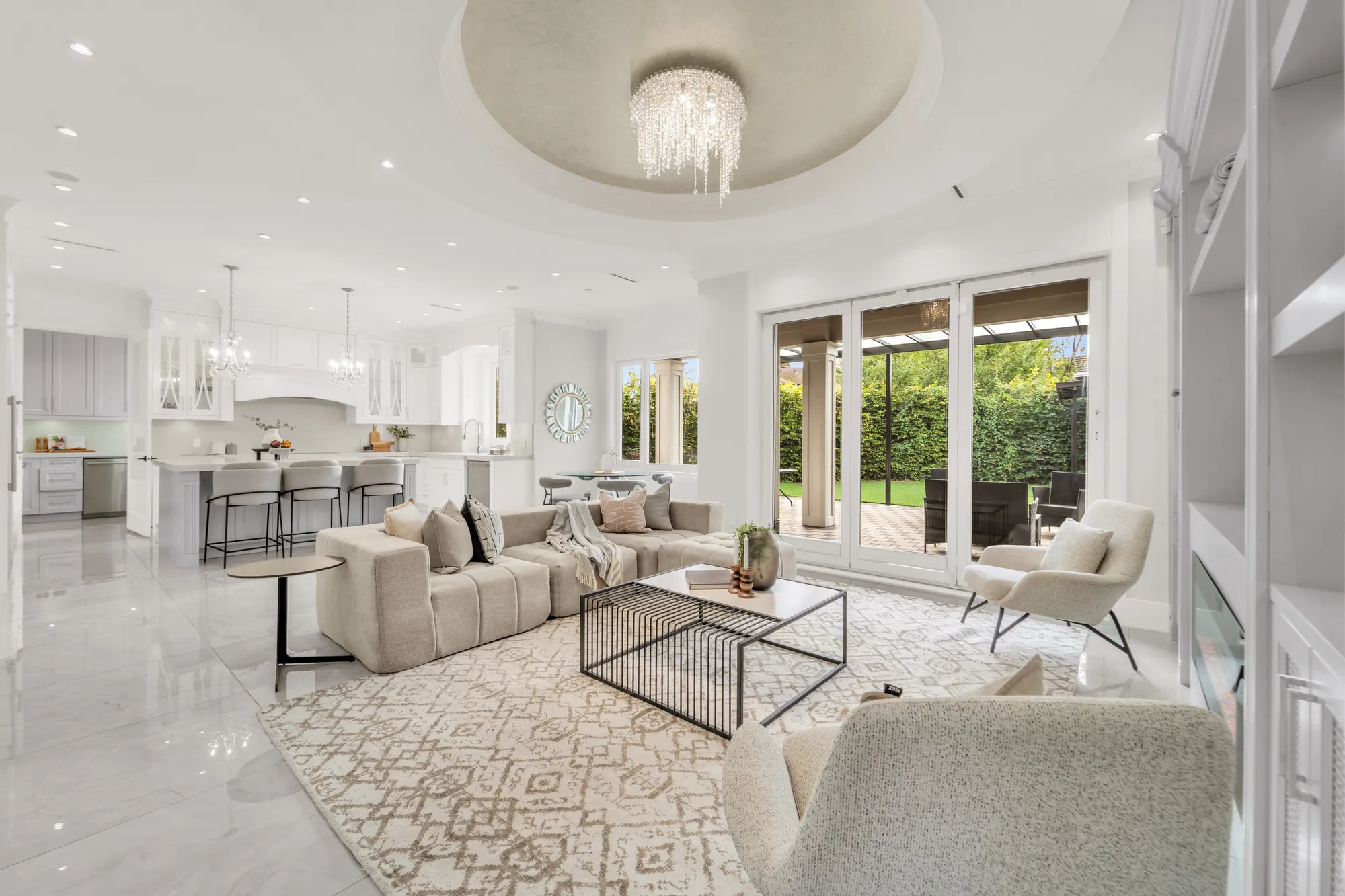 From the family room to the kitchen: the island with three stools, white cabinets and two chandeliers, glass doors to the covered patio and the yard at the right