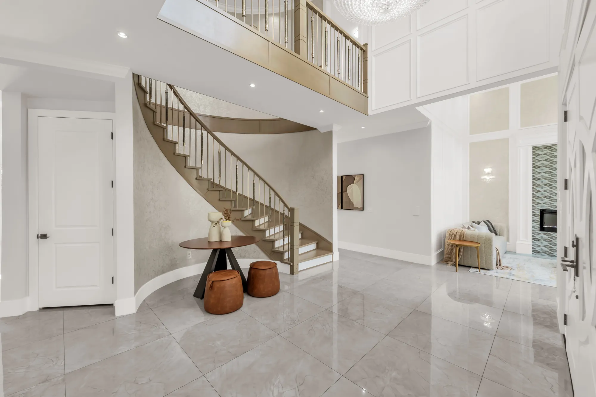 The foyer: a curved staircase with a wood handrail and metal balusters rising to an open landing, a round table and two leather stools, a crystal chandelier overhead, a large-format tiled floor