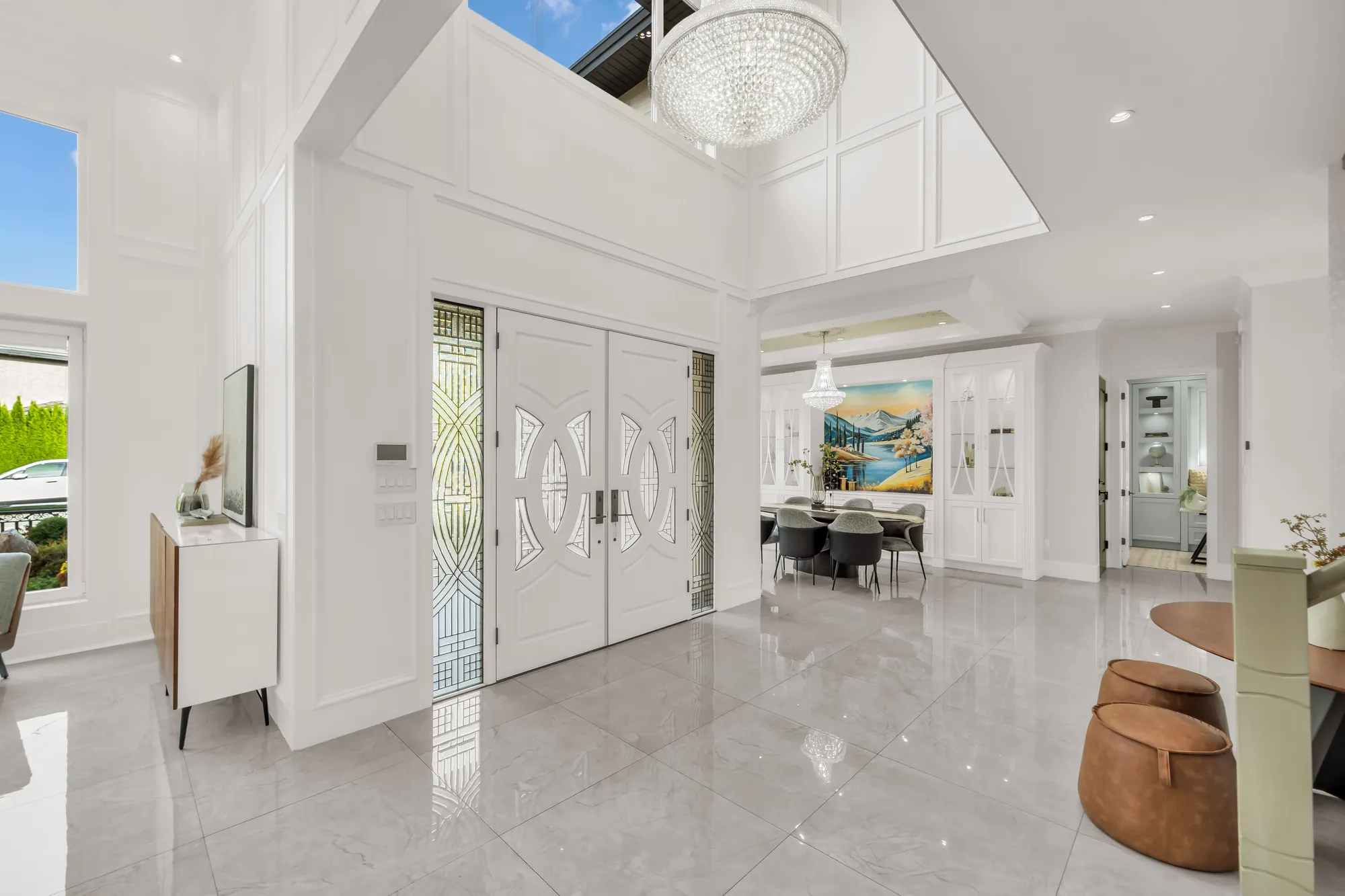 The foyer toward the double front doors: white doors with leaded-glass panels and sidelights, a crystal chandelier in the two-storey space above, the dining room at the right