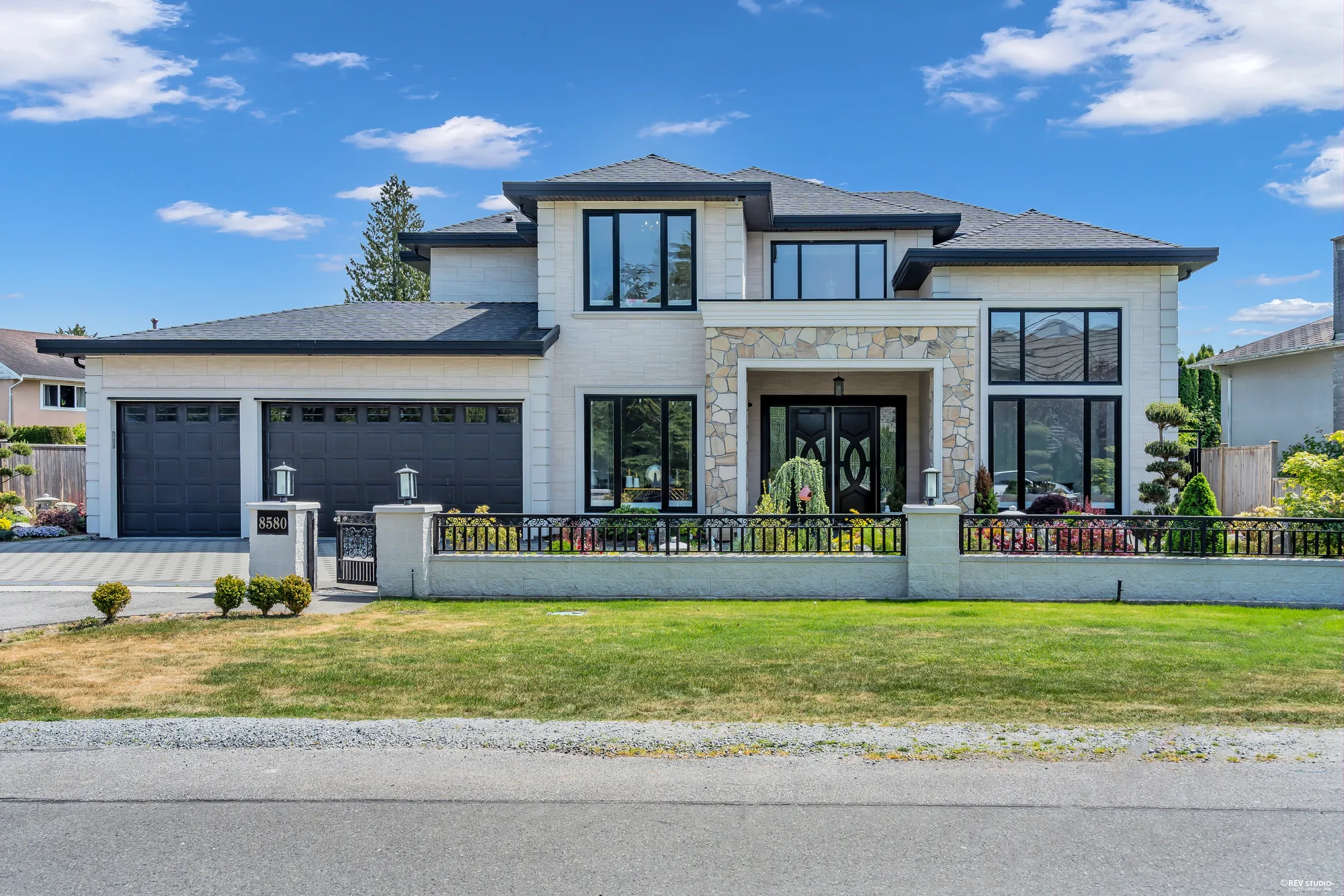 The front of the house from the street: two storeys in white cladding with a stone-faced entry and black-framed windows, a single and a double garage door at the left, a low white wall with a black iron railing and lamp posts, the number 8580 on the gate post, a lawn in front