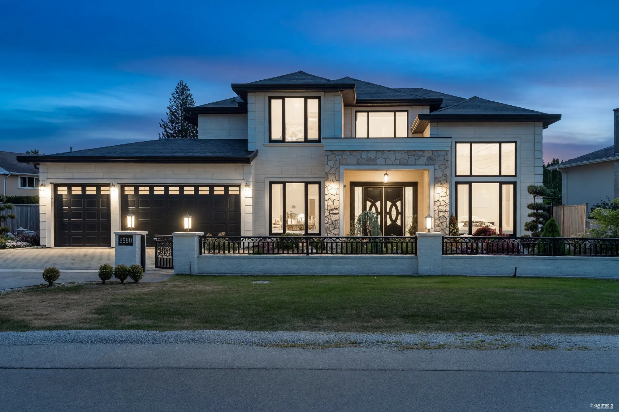 The front of 8580 Camden Crescent at dusk: two storeys in white cladding with a stone-faced entry and black-framed windows, the windows lit, a low white wall with an iron railing and lamp posts, a lawn in front