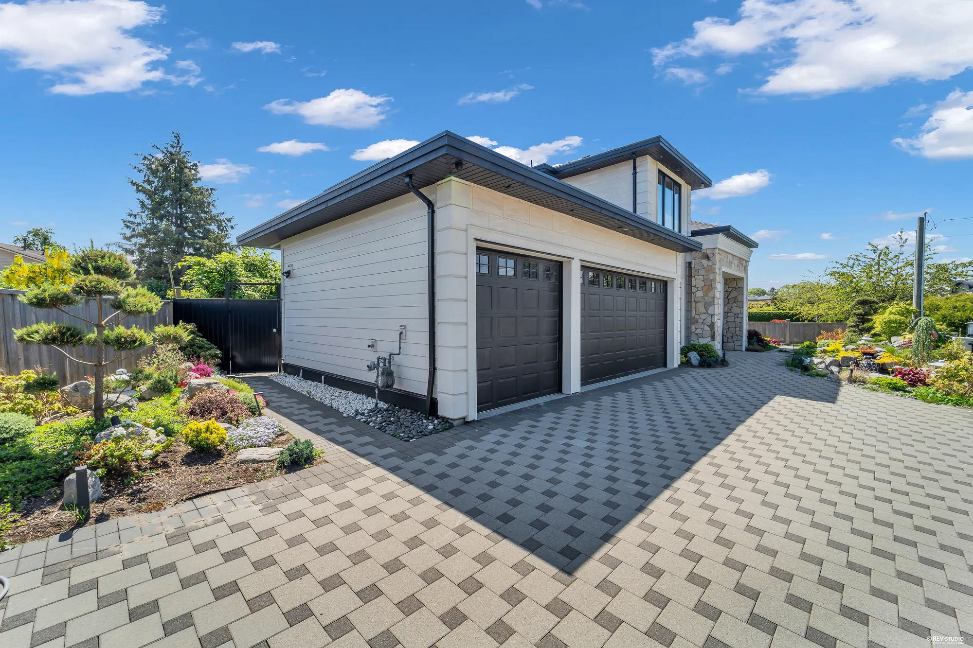 The garage doors from the side of the drive: two dark panelled doors in white siding, a paved court in grey pavers, clipped pines and planting at the left