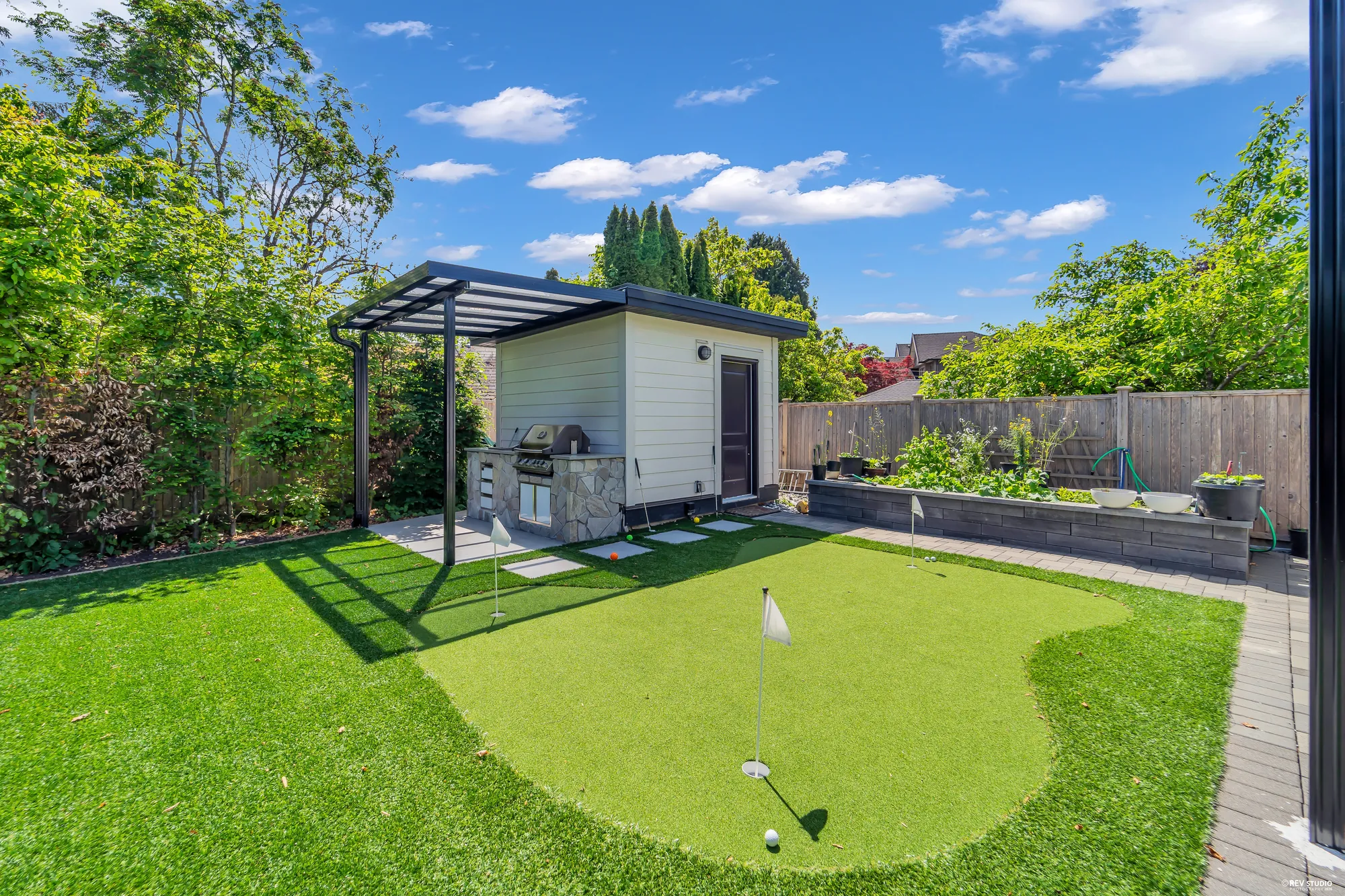 The putting green at the back of the yard: a flag on the green, a garden shed with a dark door, a grill set in a stone counter under a canopy beside it, raised planting beds along the back fence