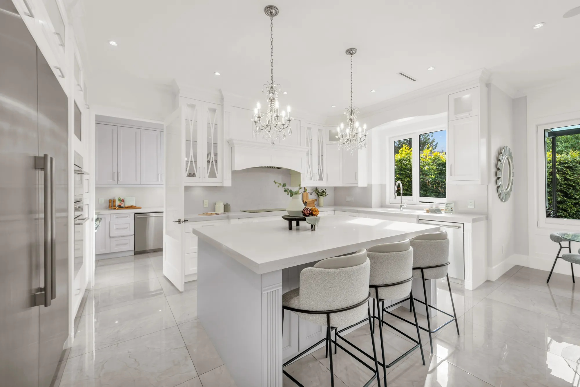 The kitchen: a large island with three upholstered stools, two crystal chandeliers, white shaker cabinets with glass-front uppers, a stainless built-in refrigerator at the left, a window over the sink
