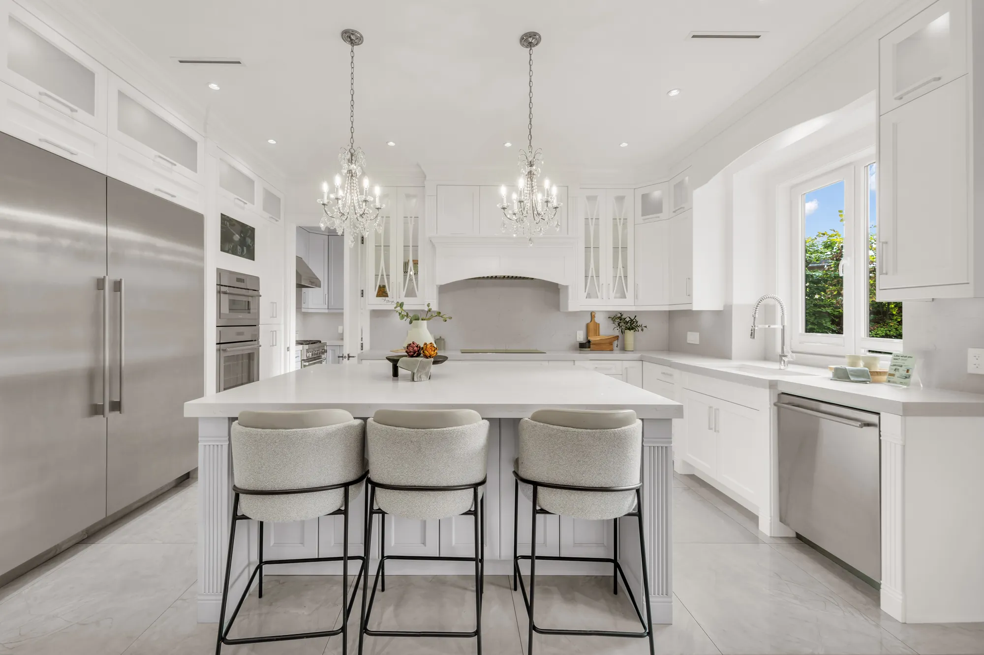 The kitchen toward the stainless refrigerator and freezer columns and the built-in ovens, the white hood surround, a dishwasher and the sink under a window