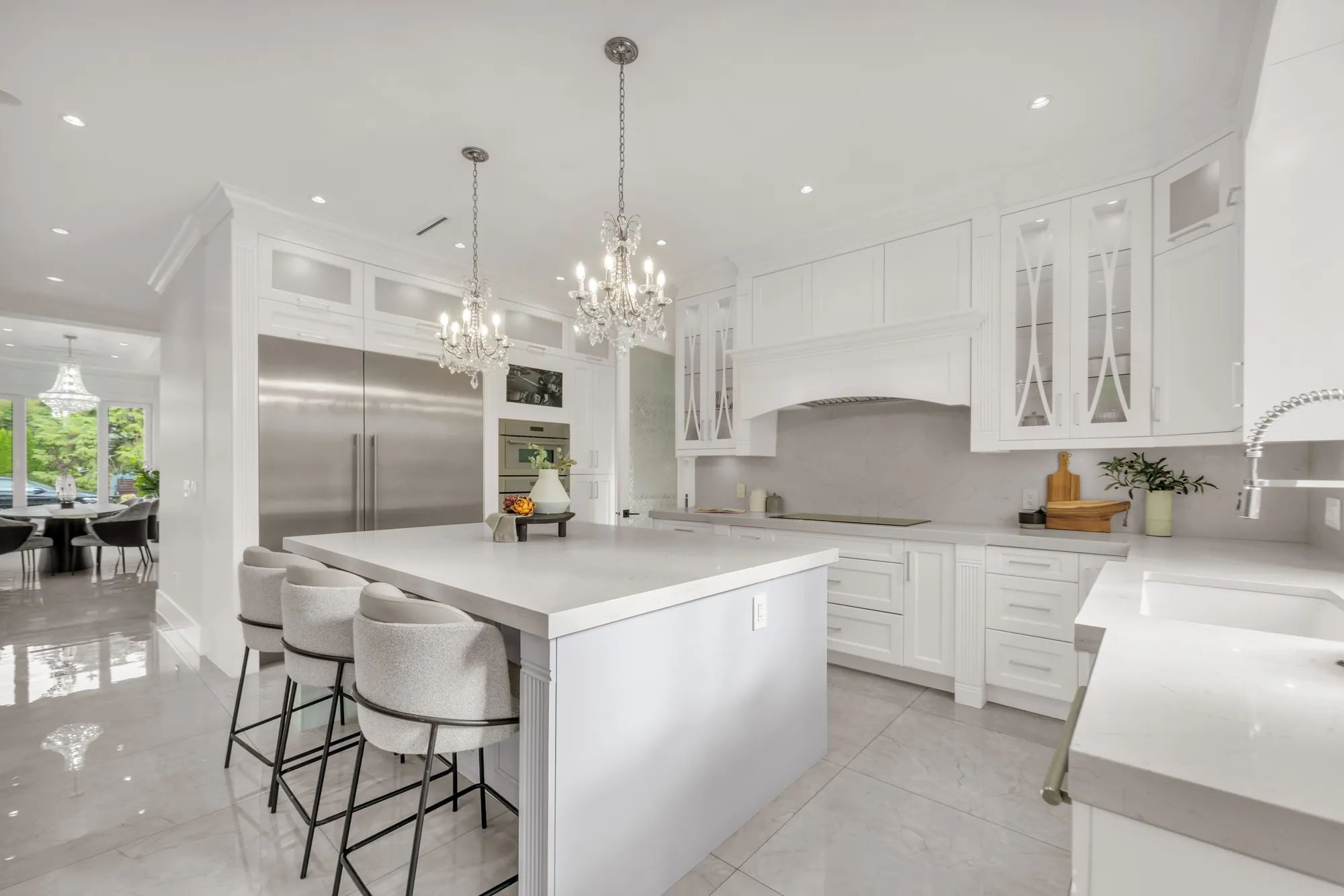 The island from its end, its stools, the refrigerator columns and the hood surround, the dining room and its chandelier beyond at the left