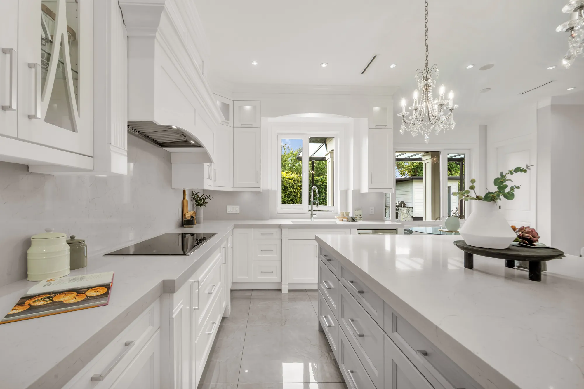 A black cooktop in the counter under a stainless hood insert, white cabinets with glass-front doors, the island at the right, a window beyond