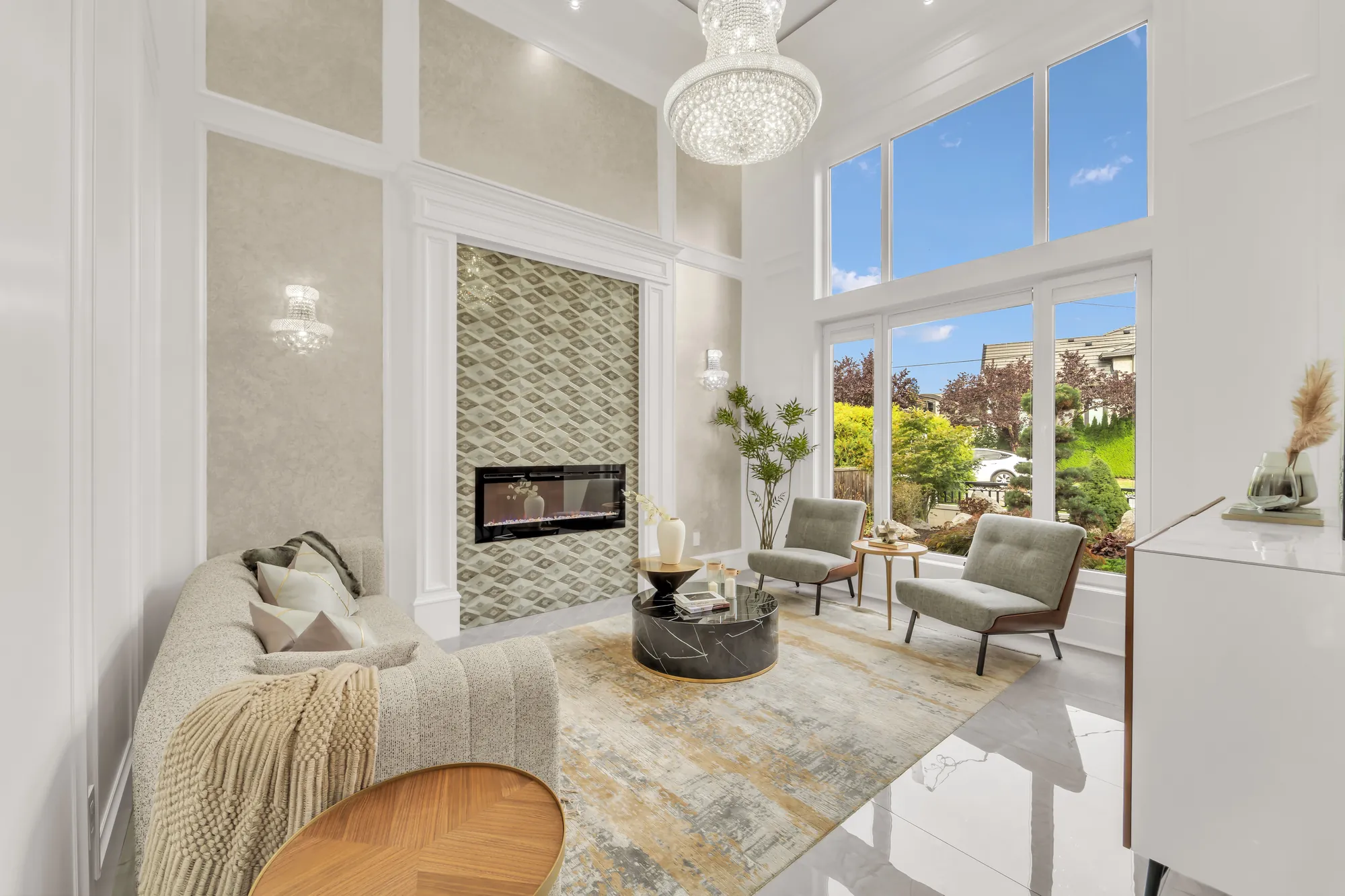 The living room: tall windows in two tiers onto the front garden, an electric fireplace in a tiled surround between wall sconces, a crystal chandelier, a curved sofa, two lounge chairs and a round table on a rug