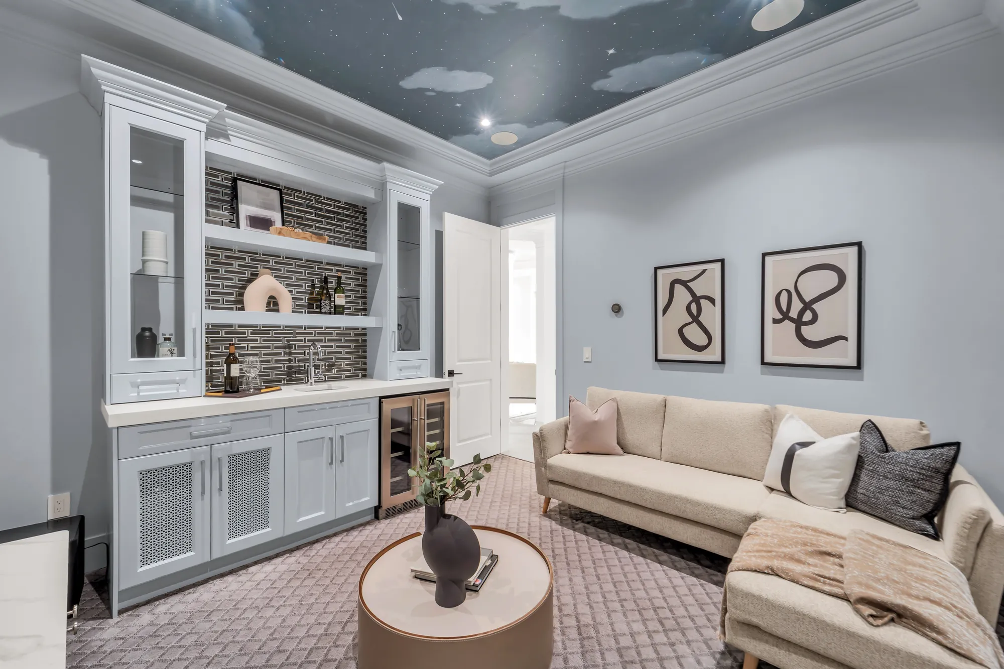 The media room: pale blue walls, a ceiling painted as a sky with clouds, a built-in bar with glass-front cabinets, open shelves and a wine fridge, a sectional sofa and a round ottoman on a patterned carpet