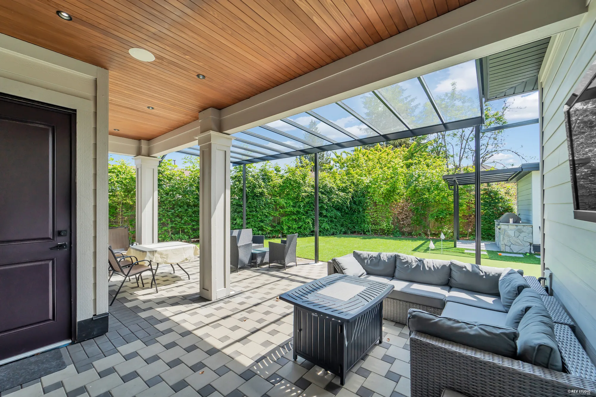 The covered patio: a wood-lined ceiling with pot lights, a glass-roofed canopy beyond it, an outdoor sectional around a fire table, the lawn and the putting green beyond, a door to the house at the left