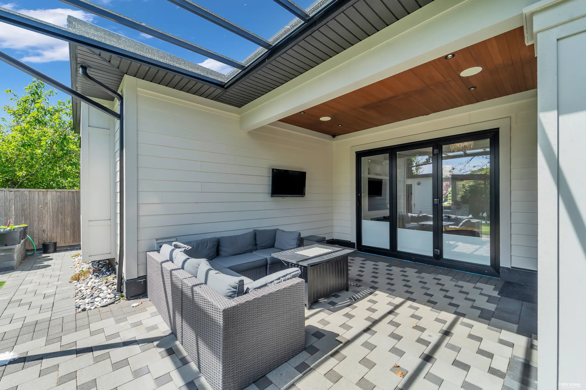 The covered patio toward the house: a wall-mounted television, sliding glass doors to the family room, the outdoor sectional and fire table on grey pavers
