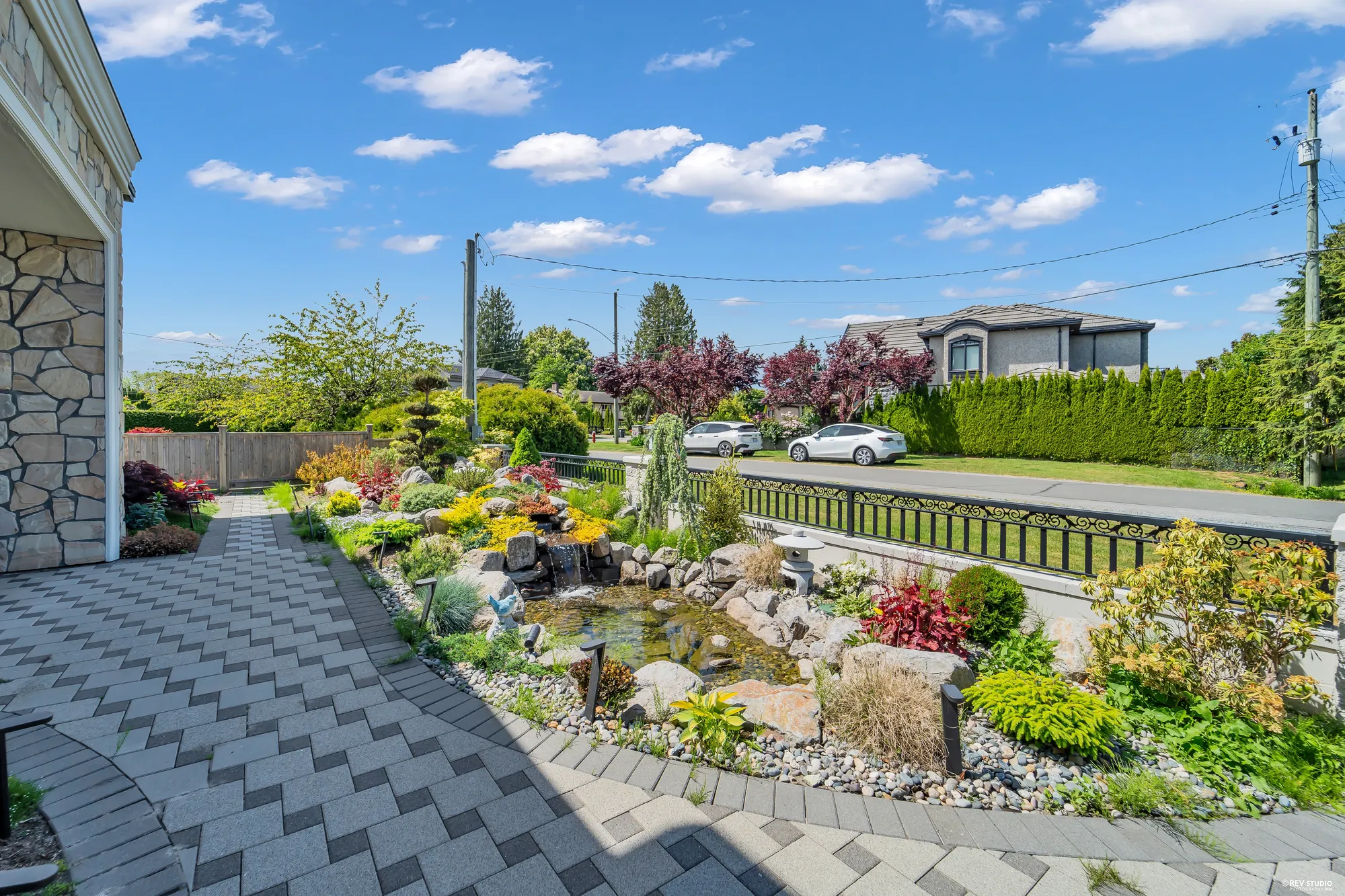 A pond in the front garden: rocks, a small waterfall and flowering planting along a paver path beside the house, the iron railing and the street beyond