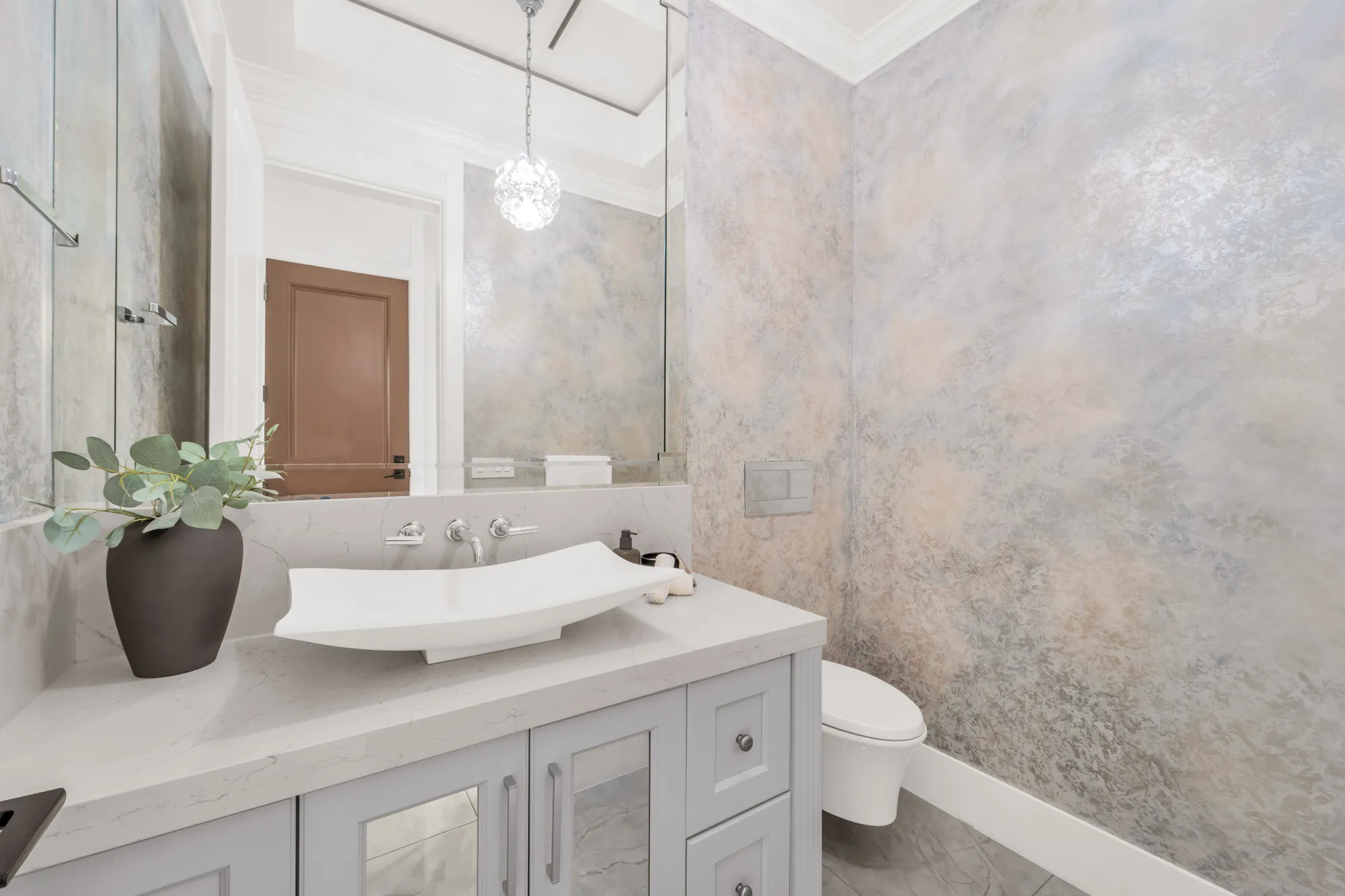 The powder room: a white stone basin on a grey vanity, a wall-mounted faucet, a wall-hung toilet, plaster-finished walls in soft grey and gold, a crystal pendant