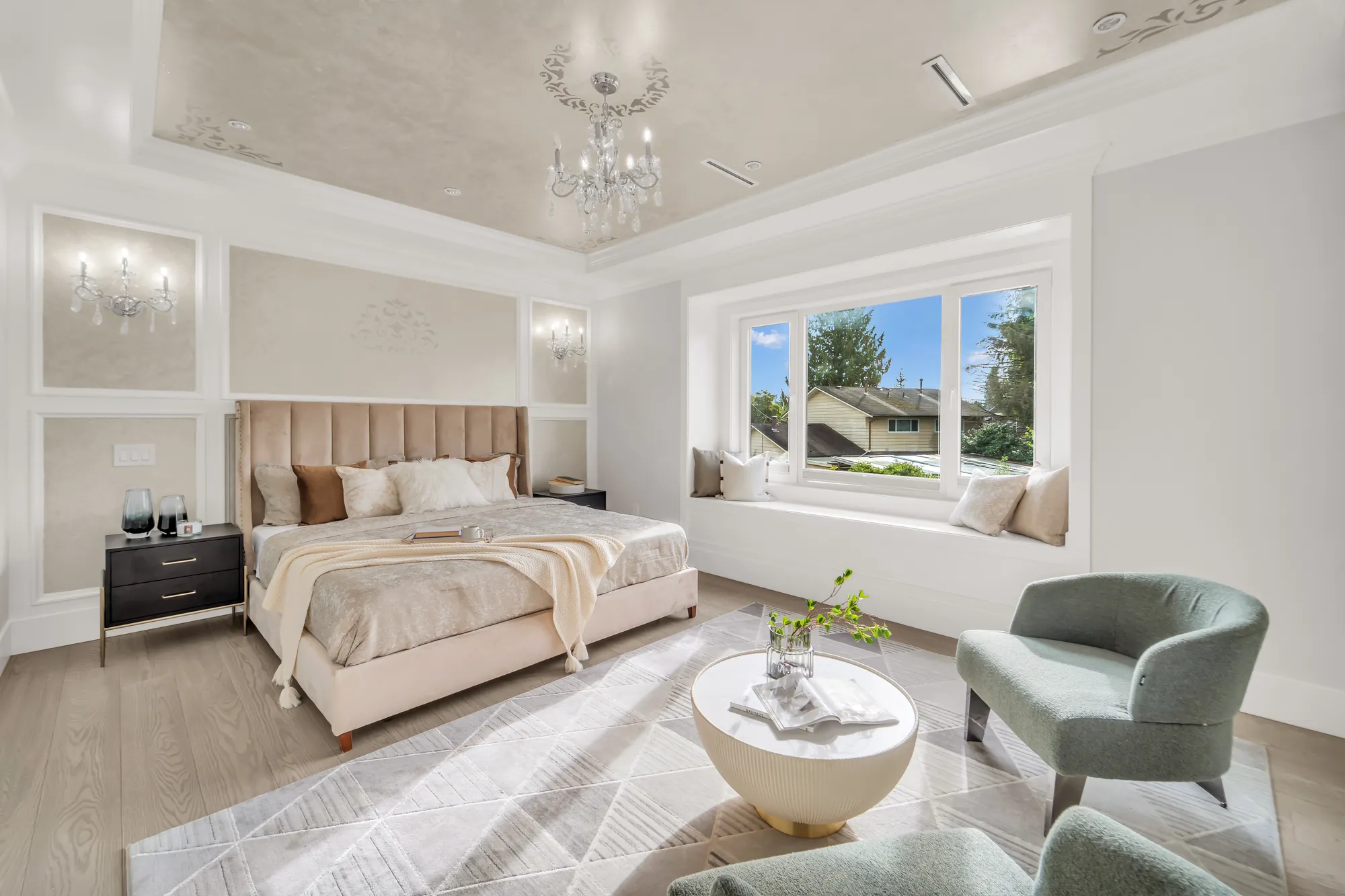 The primary bedroom: a tray ceiling with a painted panel and a crystal chandelier, an upholstered bed between wall sconces, a window seat under a wide window, two lounge chairs and a round table on a rug