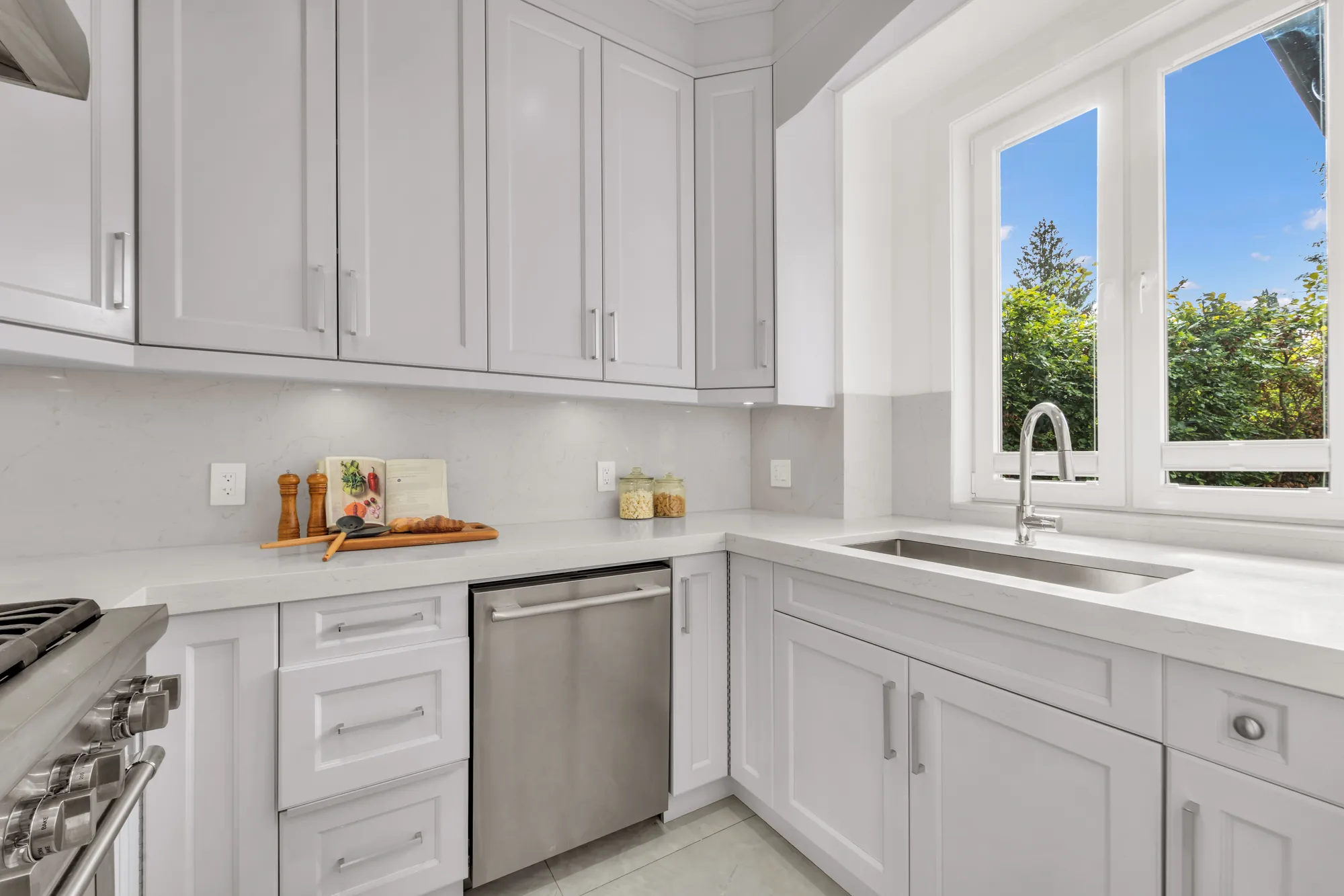 The second kitchen: white shaker cabinets on two walls, a sink under a casement window, a stainless dishwasher, white counters and backsplash