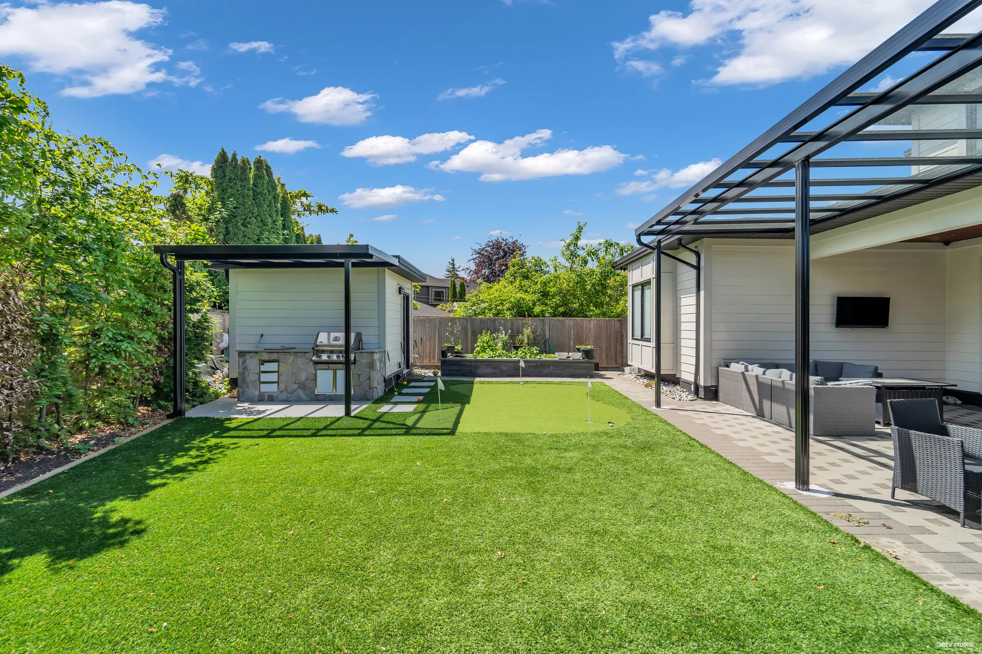 The back yard from beside the house: the lawn, the shed and the grill at the far end, a raised bed along the back fence, the covered patio with its television and furniture at the right